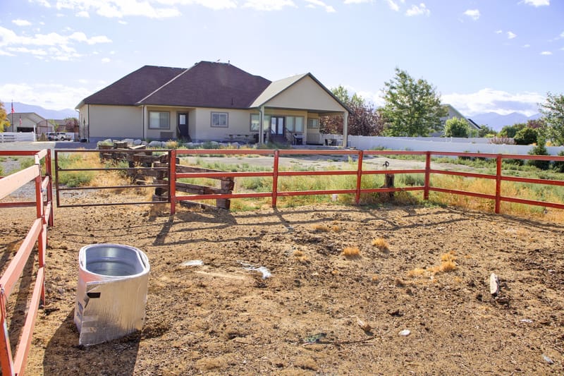 One of the three corrals at ground level, with red pipe fencing, a water trough, and the house behind it.