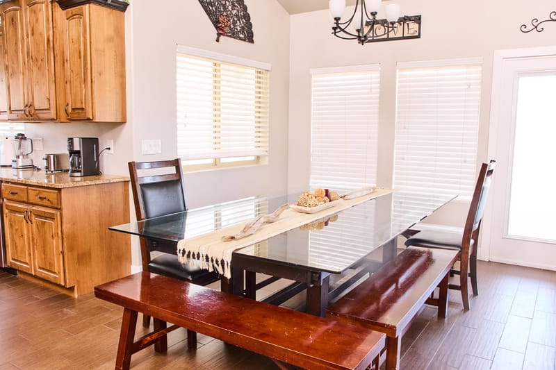 The dining room, with a long glass-topped table, a bench along one side, and a wrought-iron chandelier above.
