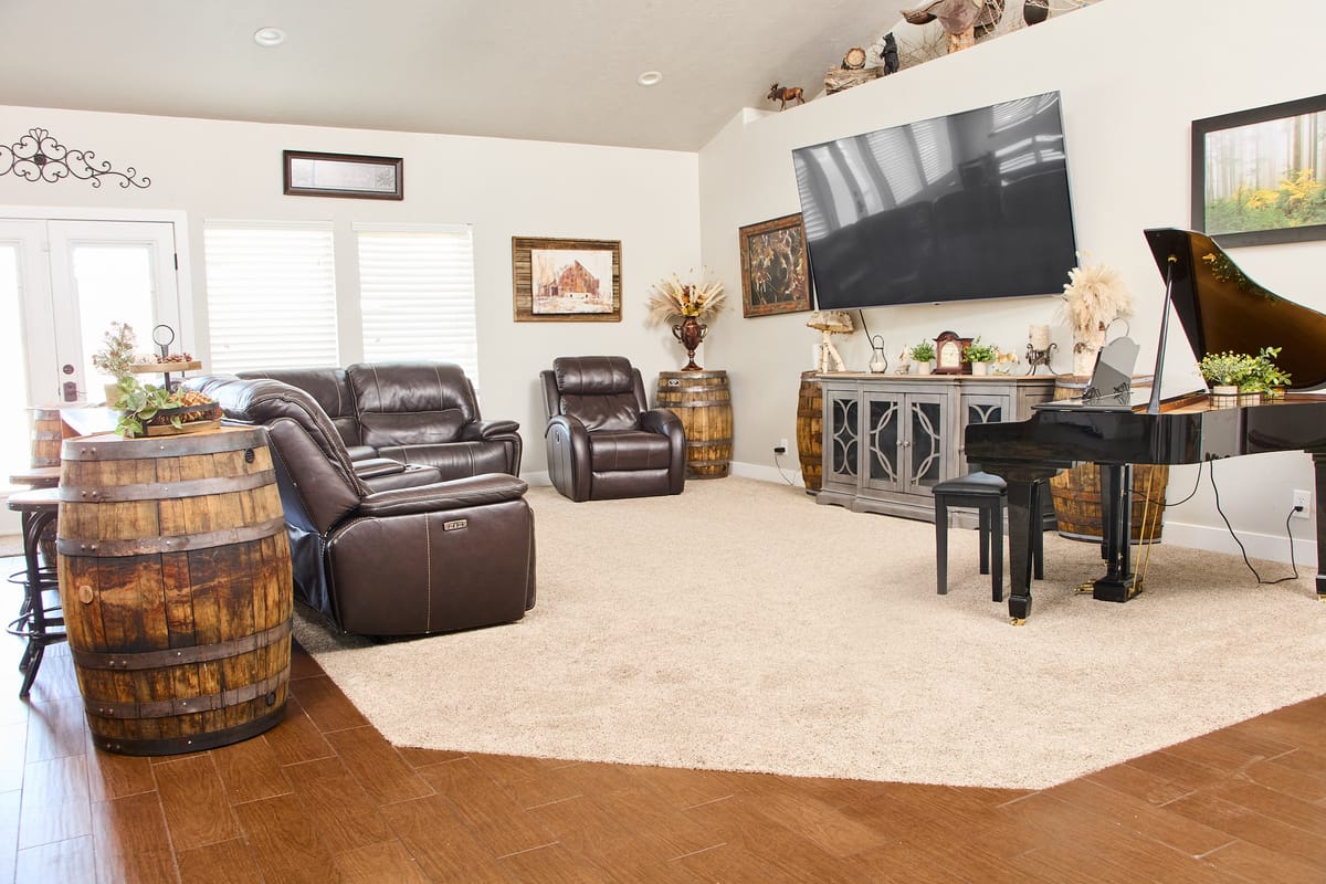 The great room, with leather reclining sofas facing a wall-mounted television, a grand piano at the far end, and a whiskey-barrel table.